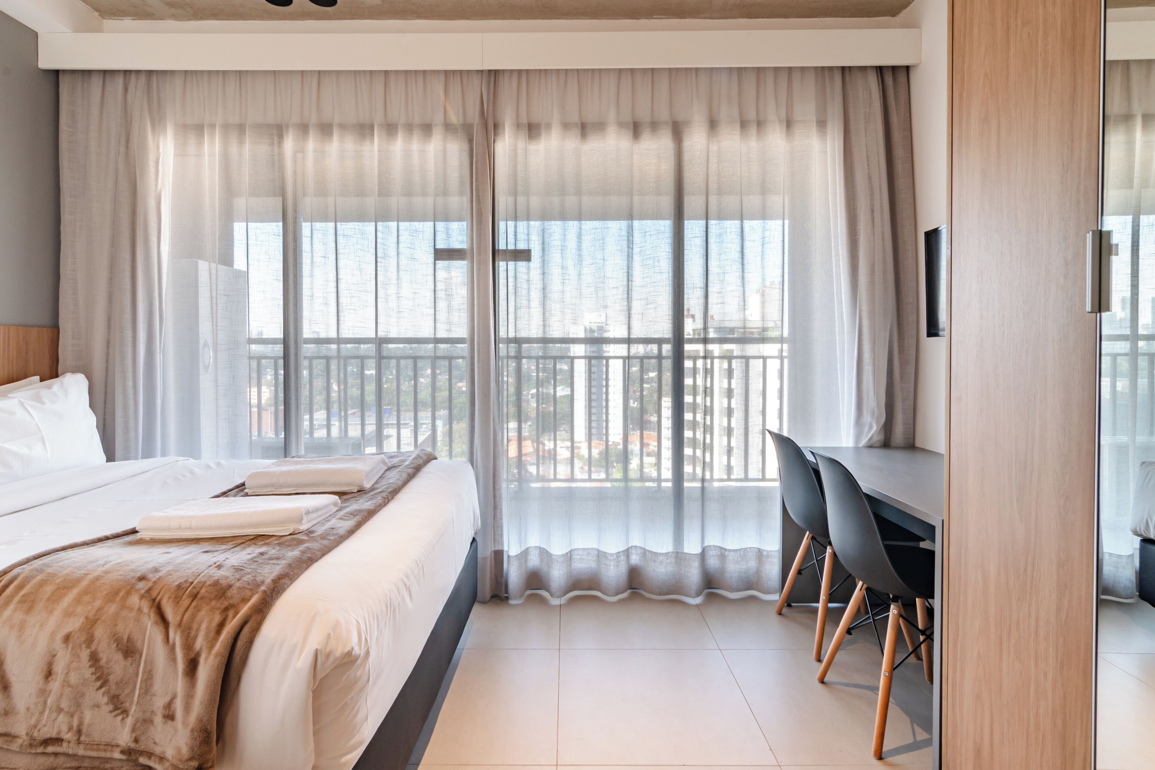 Ein modernes Hotelzimmer mit einem großen Bett, einem Schreibtisch und Blick auf die Stadt durch transparente Vorhänge.