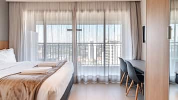 Quarto de hotel moderno com uma cama grande, uma escrivaninha e vista para a cidade através de cortinas translúcidas.