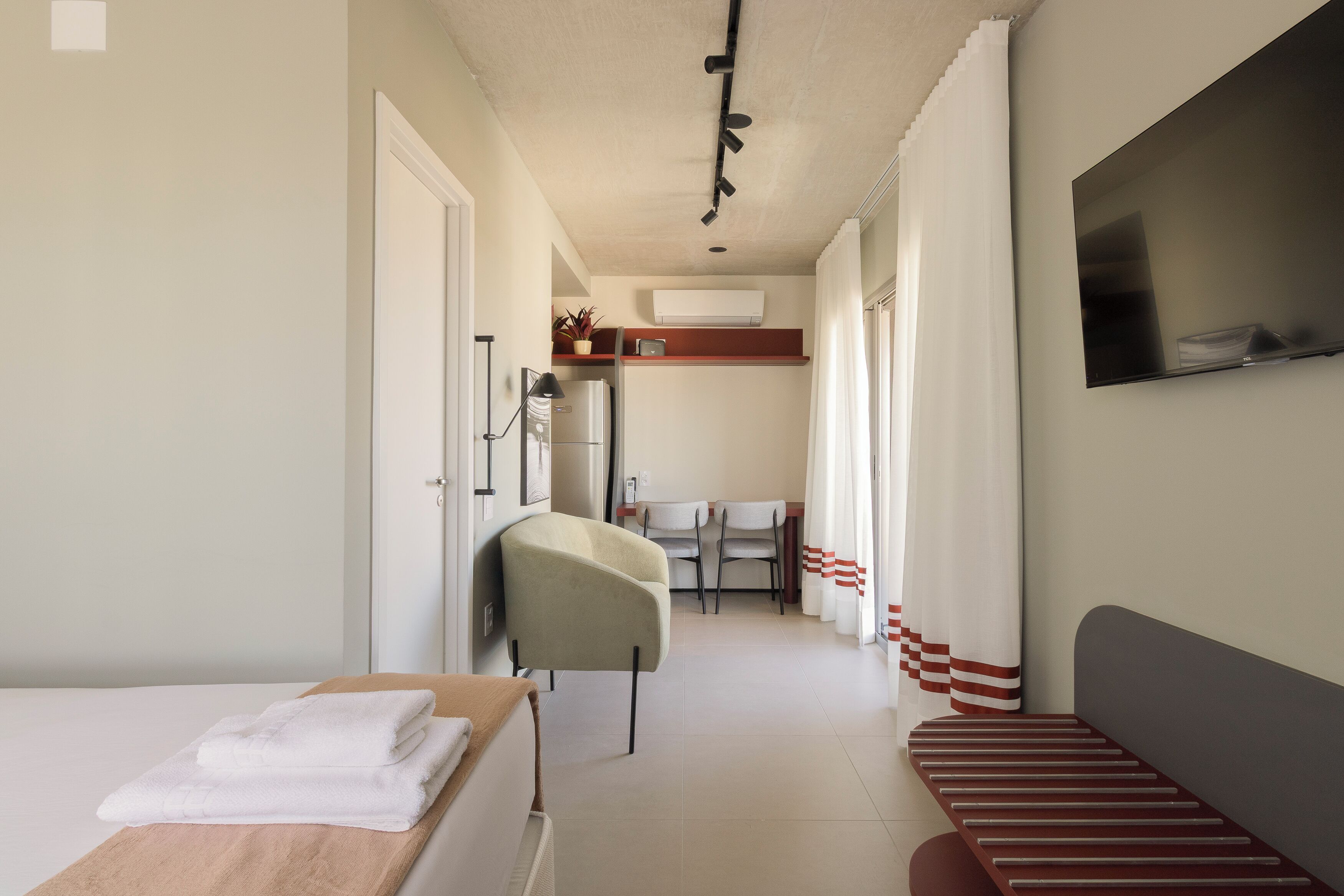 Een smalle hotelgang met een bed, een stoel, een eettafel en een televisie die aan de muur hangt.