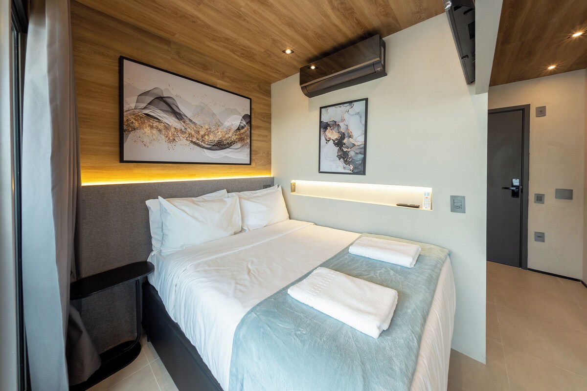 A modern hotel room with a bed, a headboard, a wall-mounted artwork, a bedside table, and a door.
