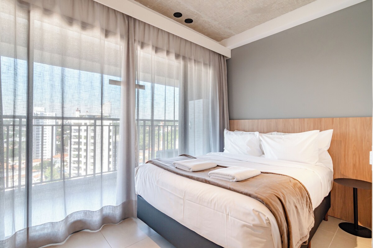 A hotel room with a large bed, a view of the city through sheer curtains, and a balcony.