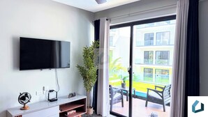 A modern living room with a flat-screen TV mounted on the wall, a small wooden cabinet, a potted plant, and a sliding glass door leading to an outdoor area with a pool and seating. - Walk 2 Fun, Beaches, Dining, Shopping, Nightlife (Punta Cana)