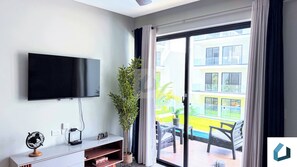 Ein modernes Wohnzimmer mit einem Wand-Fernseher, einer kleinen Holzkommode, einem Topf mit Pflanze und einer Glastür, die zu einem Au ßenbereich mit Pool und Sitzgelegenheiten führt.