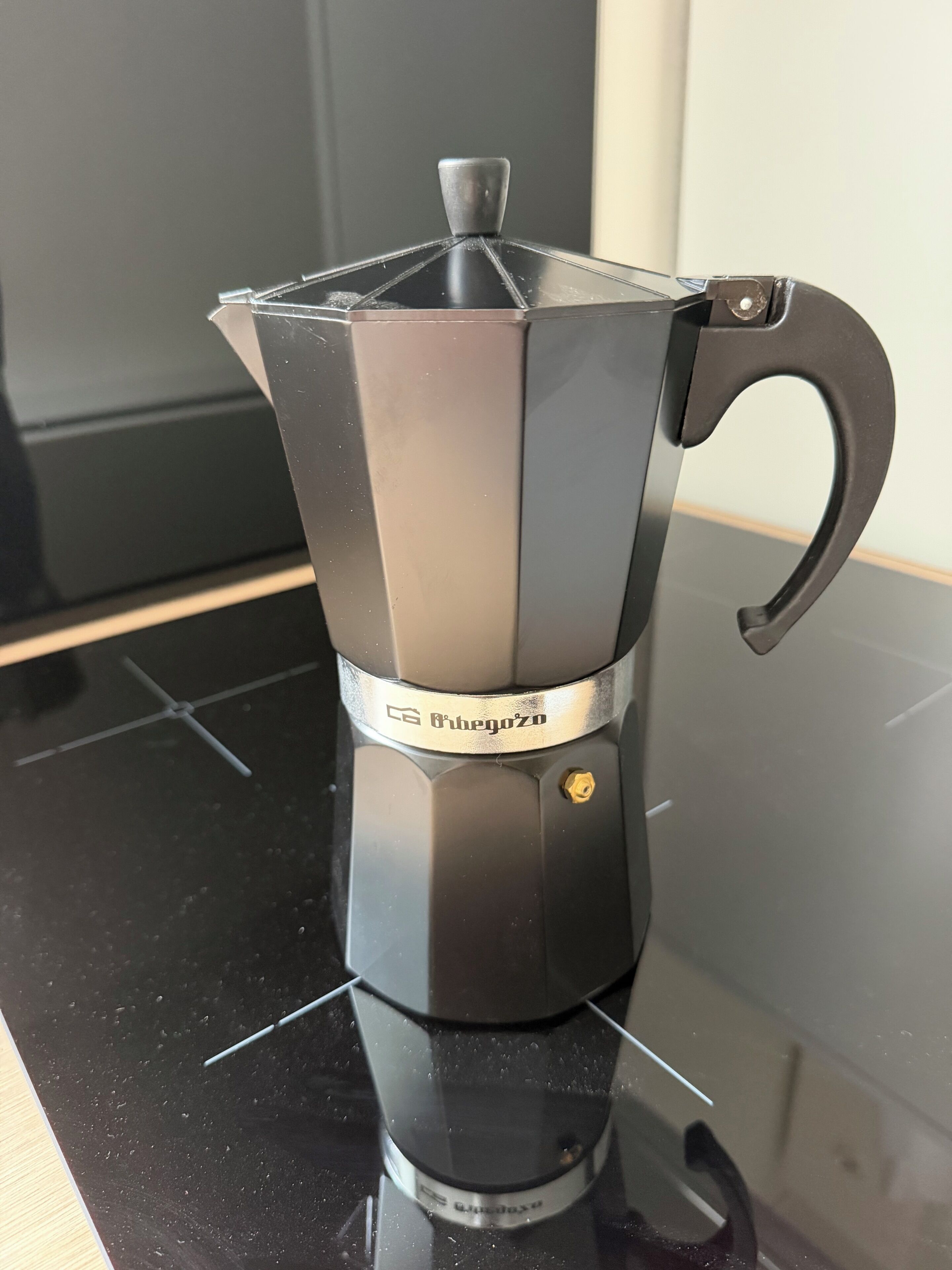A black coffee maker with a metallic band and a handle on a reflective surface.