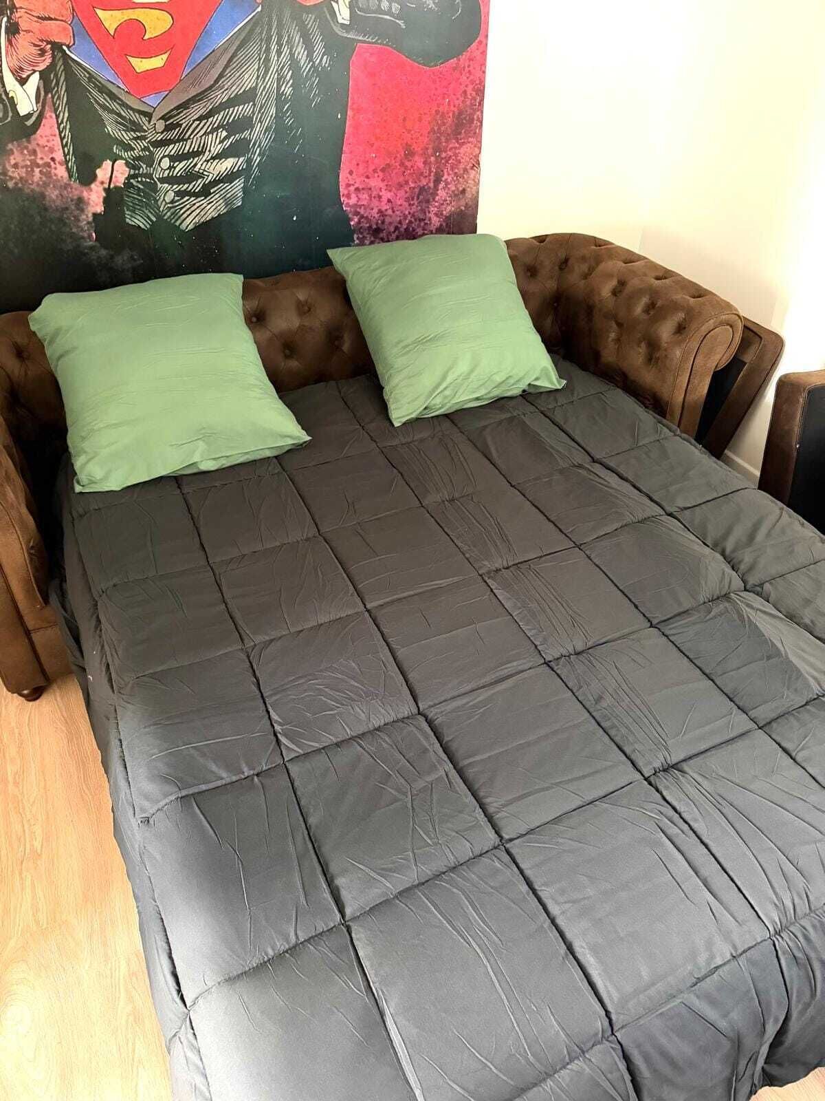 Ein Bett mit einer dunkelgrünen Decke, zwei grünen Kissen, einem braunen Kopfteil und einem farbenfrohen Poster an der Wand.