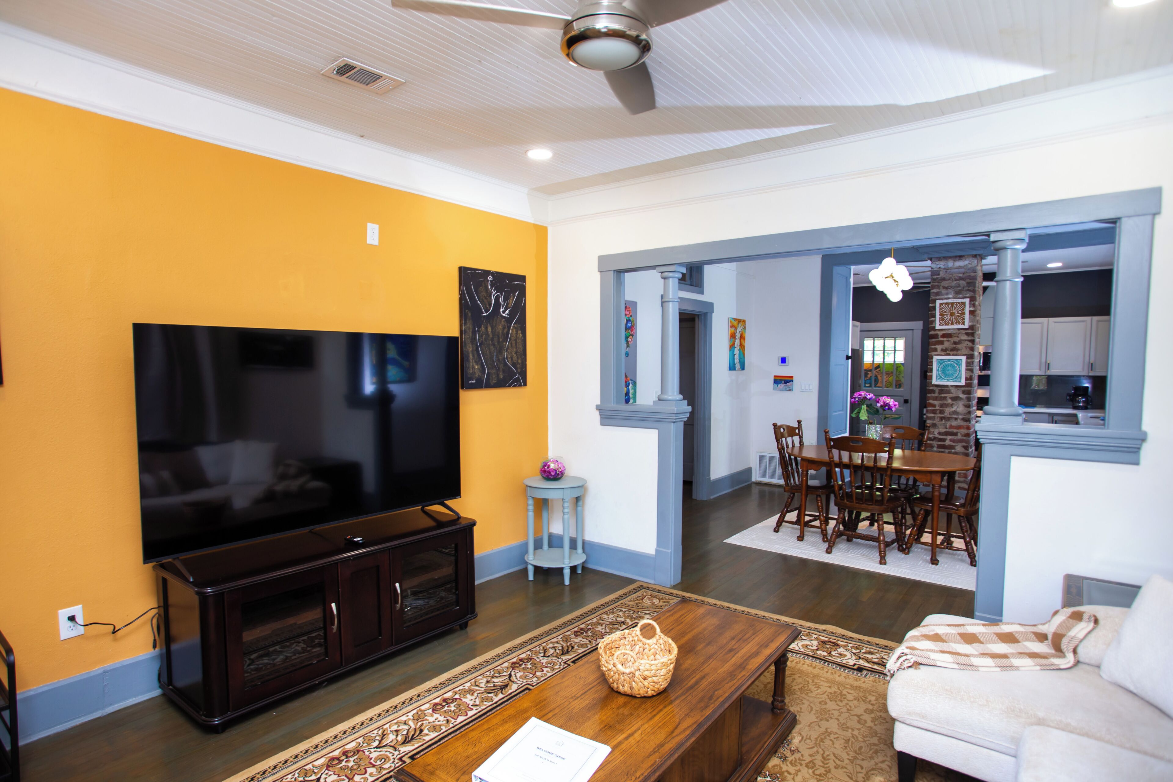 A living room with a flat-screen TV, a coffee table, and a sofa. There is a dining area with a table and chairs. The walls are painted yellow and white.