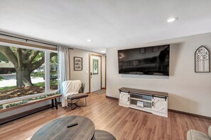 Ein Wohnzimmer mit großem Fernseher, einer Bank, einem Couchtisch und Blick auf einen Baum.