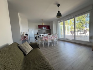 A modern living room with a sofa, a dining area with a table and chairs, a kitchen with appliances, and large windows offering a view. - 2-bedroom apartment with balcony, close to the sea (Cabourg)