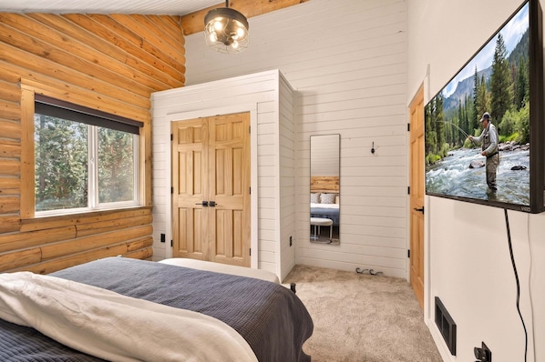 A bedroom with a bed, wooden walls, a large window, a wardrobe, and a TV displaying a nature scene.