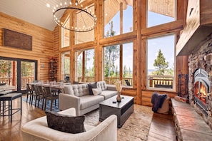 A log cabin interior with a fireplace, large windows, a dining area, and a cozy seating arrangement.