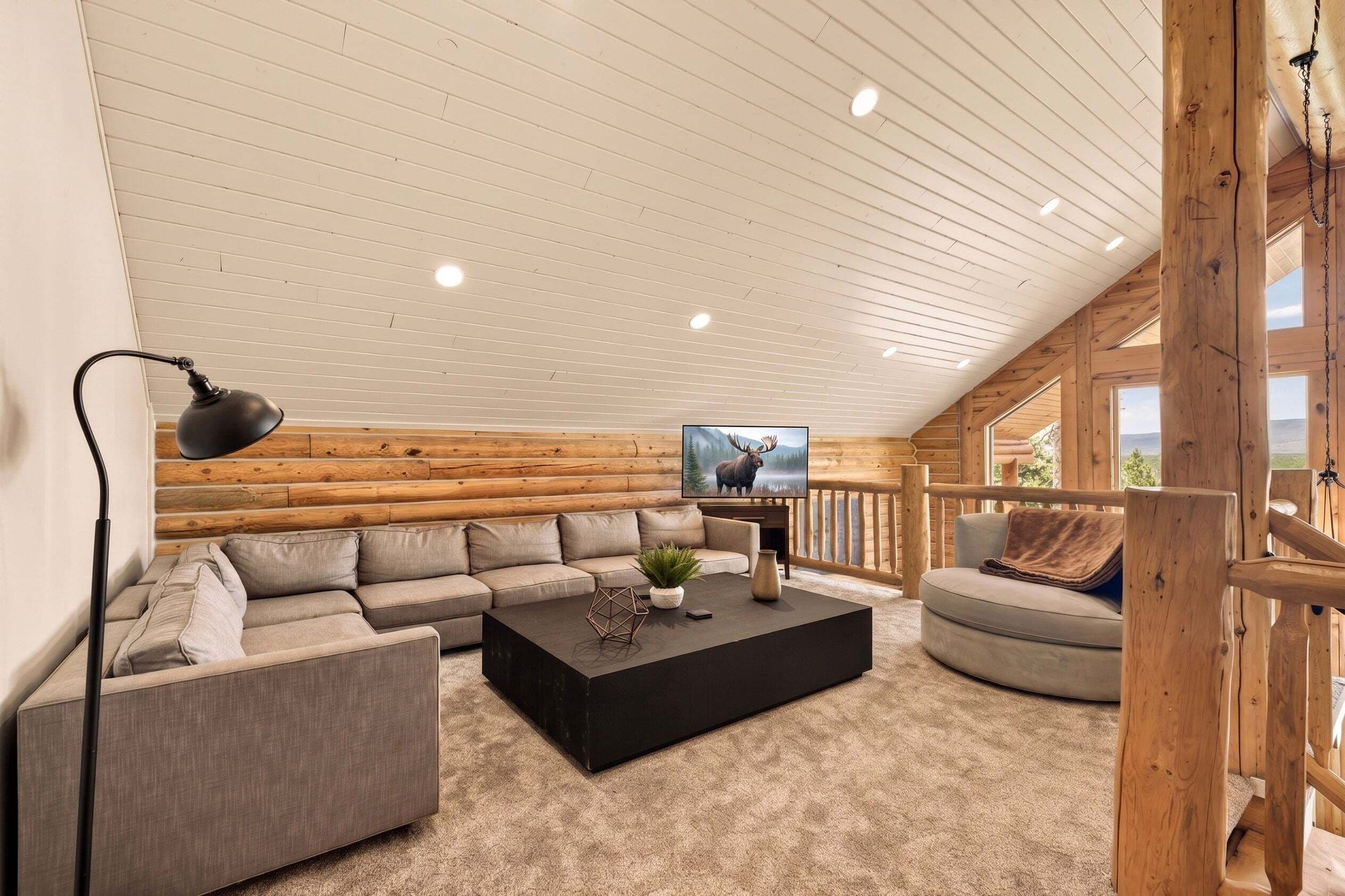A cozy log cabin interior with a sectional sofa, a black coffee table, a TV, and a wooden railing.
