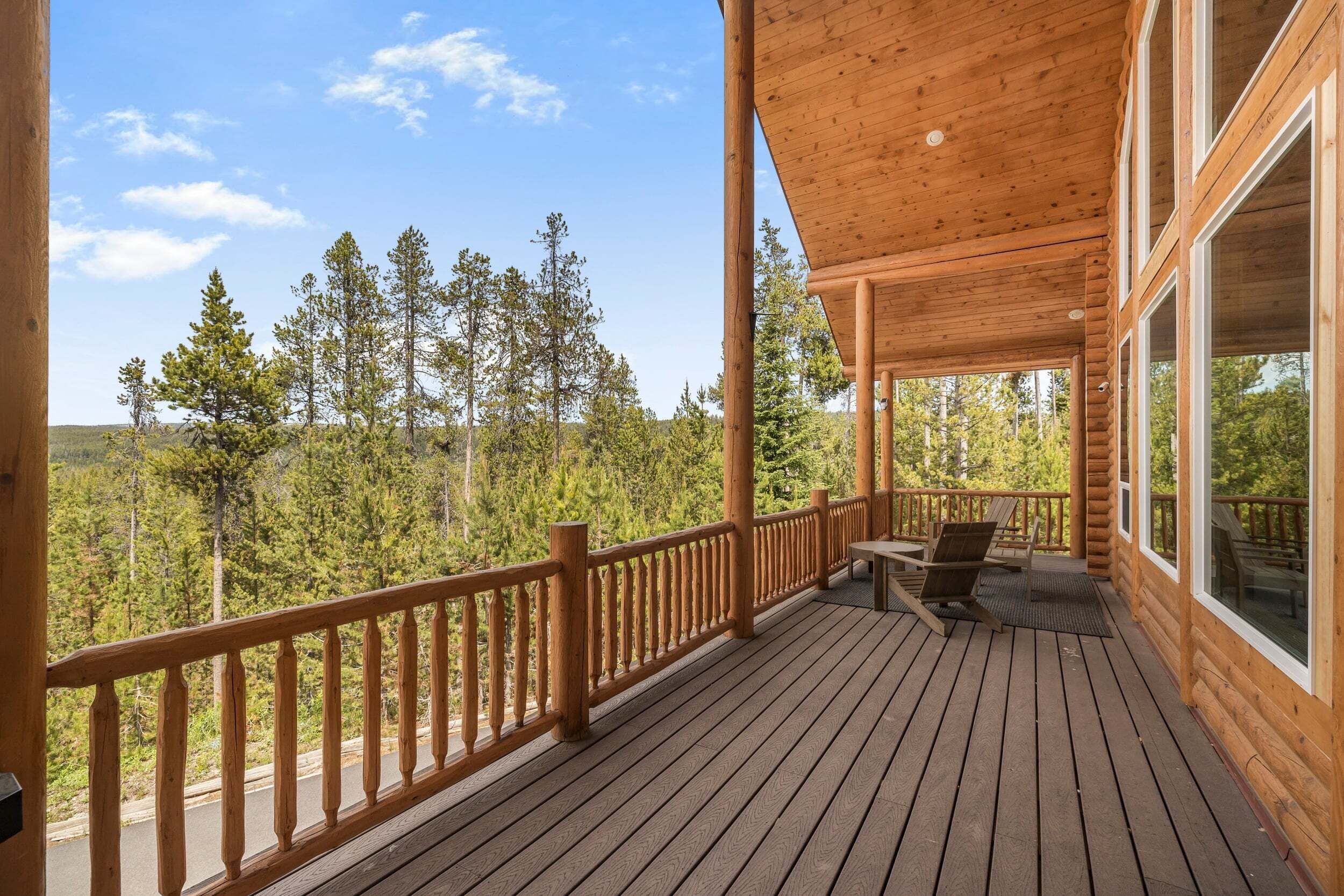 A wooden deck with a railing, a table, and chairs, overlooking a forested area.