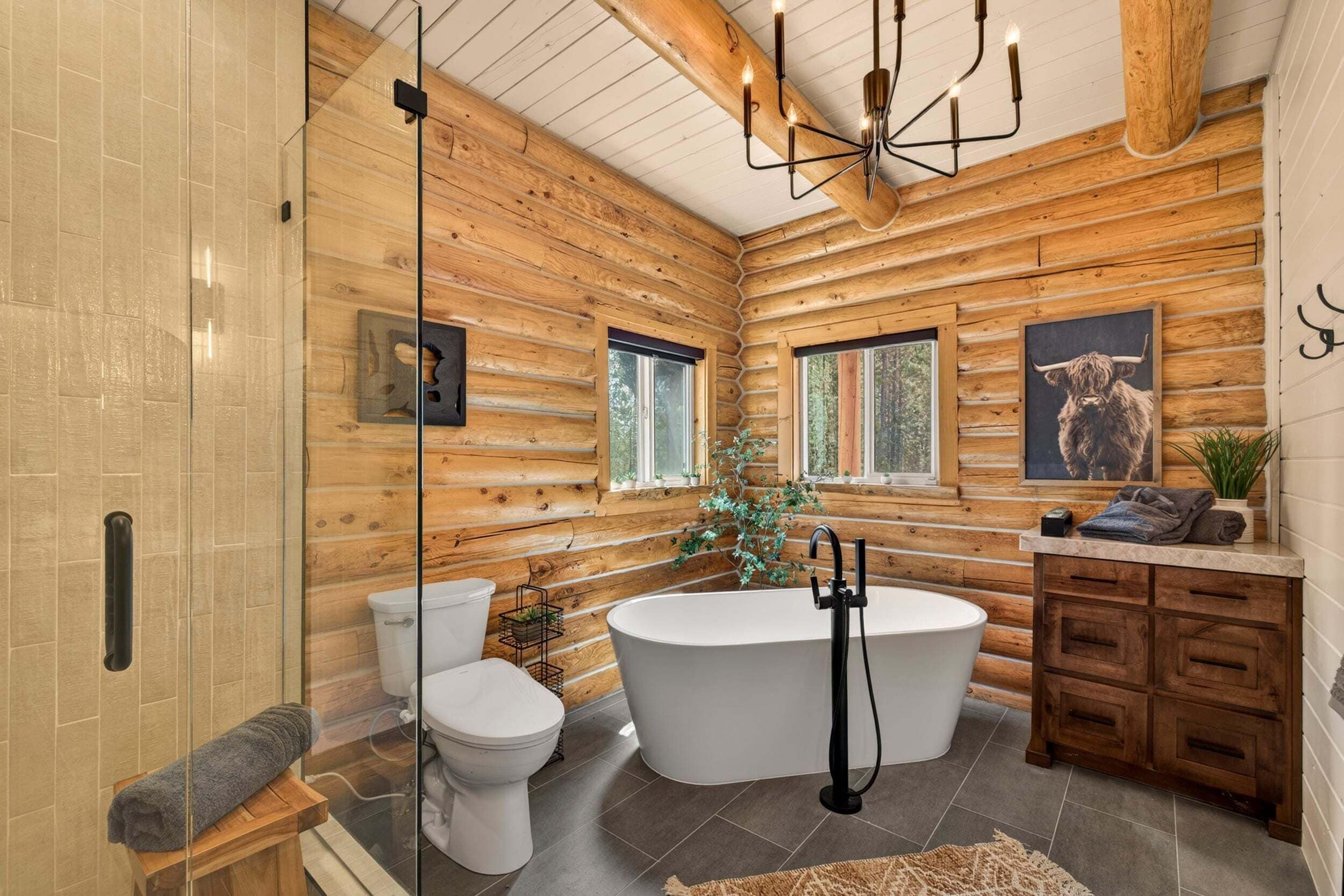 A log cabin bathroom with a freestanding bathtub, a glass shower enclosure, wooden cabinets, and a large window.