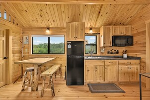 Una cocina estilo cabaña con paredes de madera, un refrigerador negro, una mesa de comedor de madera y una ventana con vistas a la vegetación.