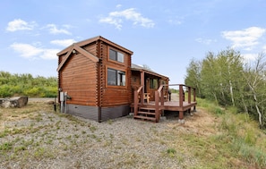 A wooden cabin with a deck, a small porch, and a gravel ground. - The Hatch Cabin (Ashton)