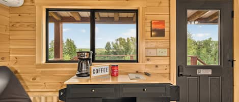 A wooden cabin interior with a 'WELCOME' sign, a coffee station, a 'NO SMOKING' sign, and a view of the outdoors.