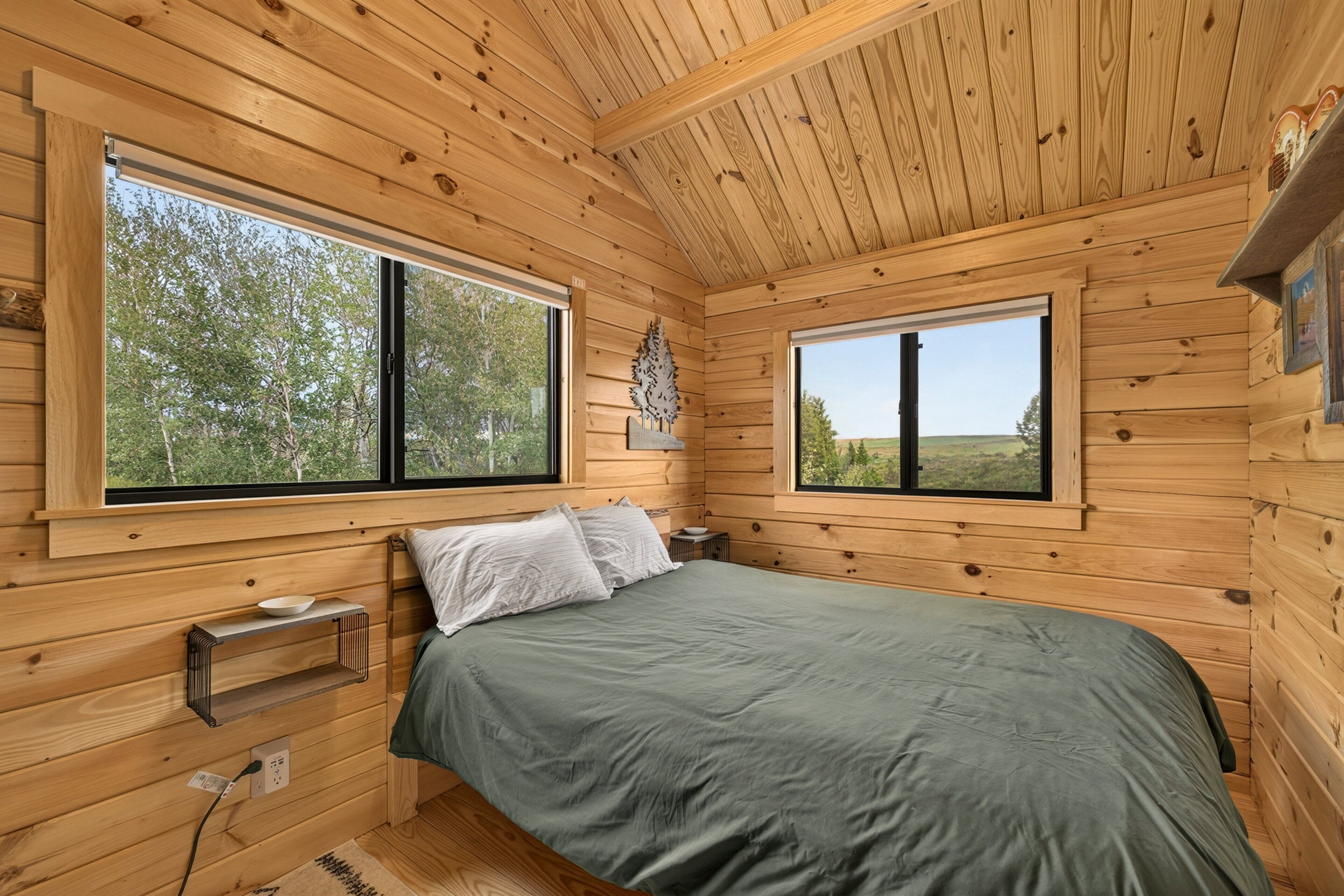 A wooden cabin bedroom with a bed, two windows, a shelf, and a power outlet.