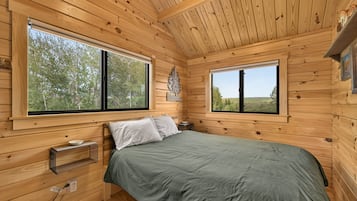 A wooden cabin bedroom with a bed, two windows, a shelf, and a power outlet.