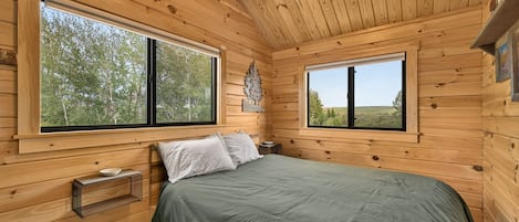 A wooden cabin bedroom with a bed, two windows, a shelf, and a power outlet.
