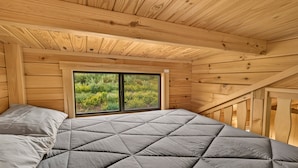 A wooden room with a bed, a window, and a view of greenery. - The Hatch Cabin (Ashton)
