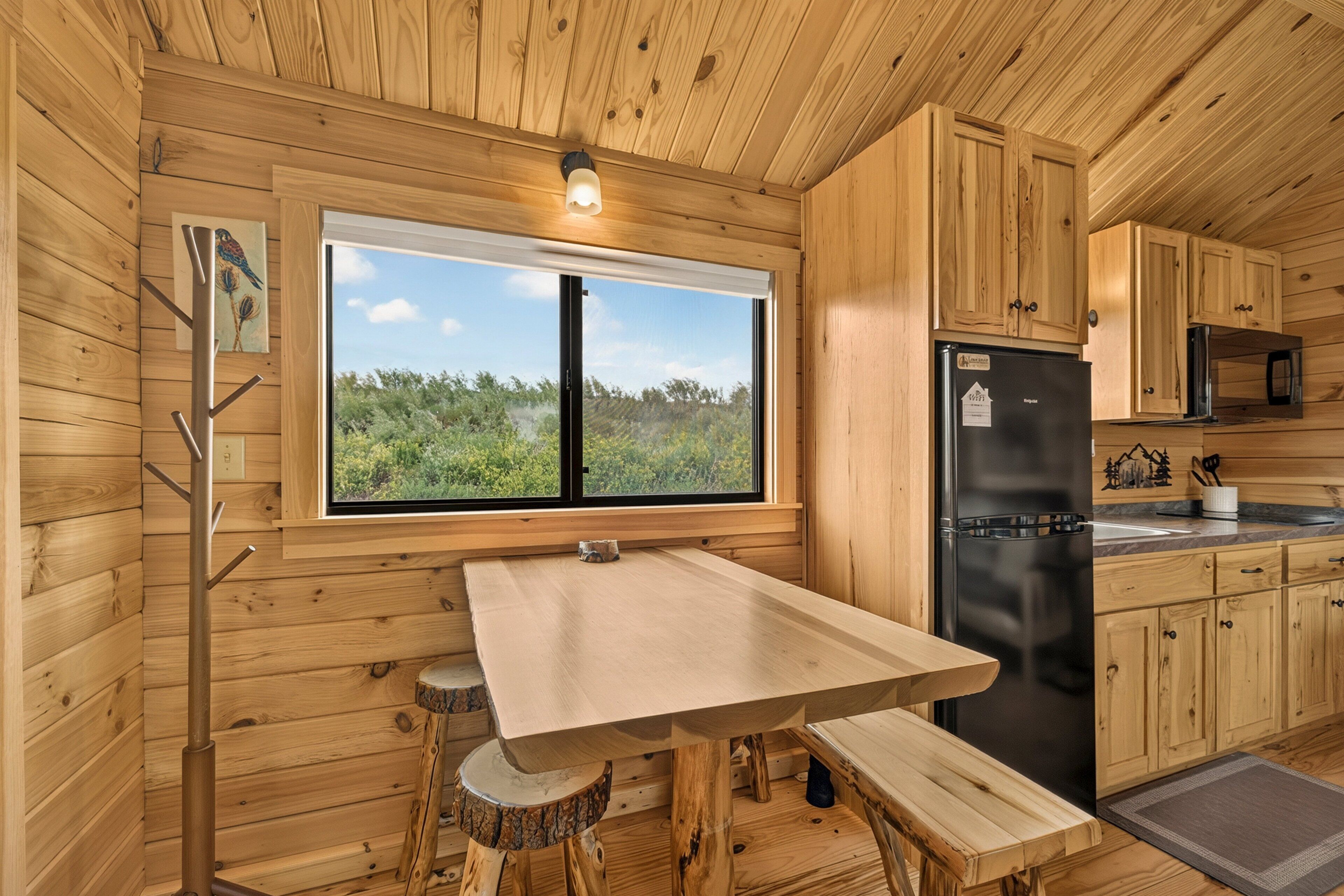 A wooden cabin interior with a dining table, a refrigerator, and a window overlooking greenery.