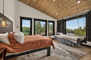 Ein Schlafzimmer mit einem Bett, einem Hängesessel und großen Fenstern mit Blick auf Grünflächen.