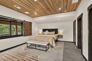 Un dormitorio con techo de madera, una cama grande, un banco y vistas a árboles por una ventana.