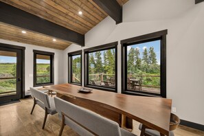 A modern interior with a wooden dining table, grey upholstered chairs, and large windows offering a view of the outdoors. - Fall River Lodge (Ashton)