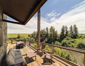 Una terraza de madera con mobiliario exterior, un muro de piedra y vistas a un río y bosque.