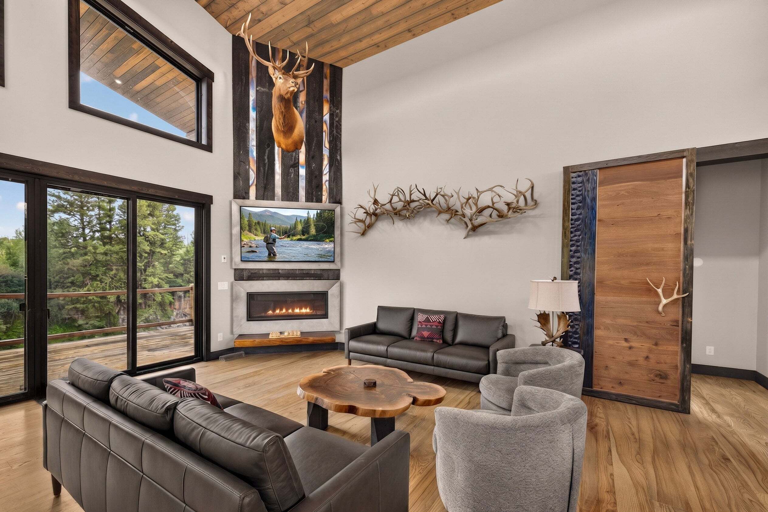A modern living room with a fireplace, leather sofas, a coffee table, and a large window overlooking a forest.