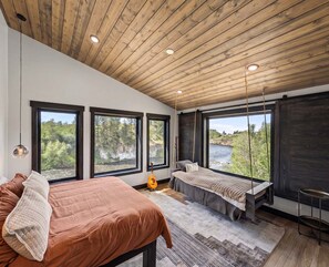 A bedroom with a bed, a bench, and large windows overlooking a river. - Fall River Lodge (Ashton)