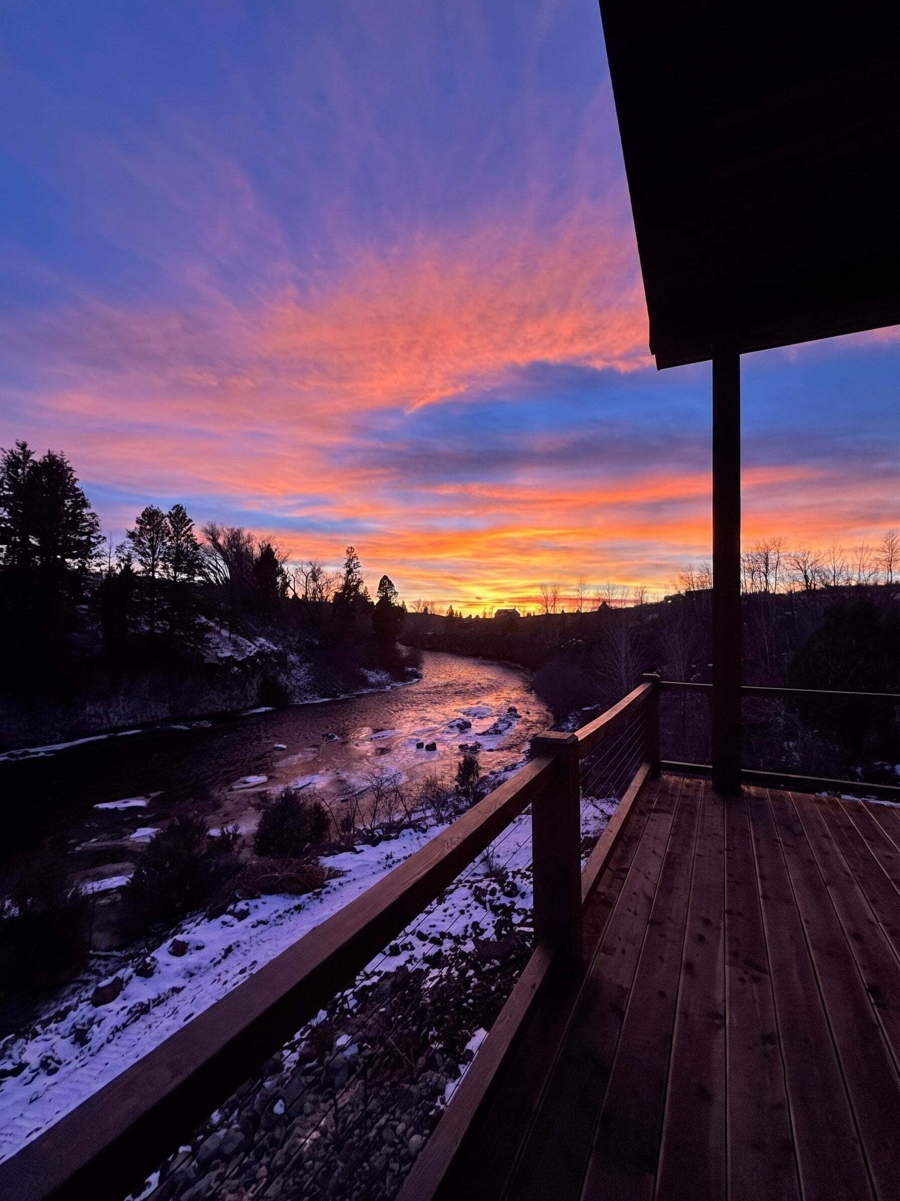 A wooden deck overlooking a river with snow on the ground and trees in the background.