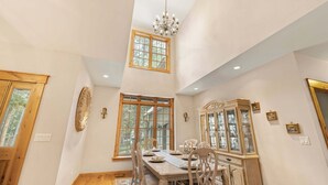 A dining room with a high ceiling, a chandelier, a large window, and a dining table set for four. - Alpine Ascent (Alpine)