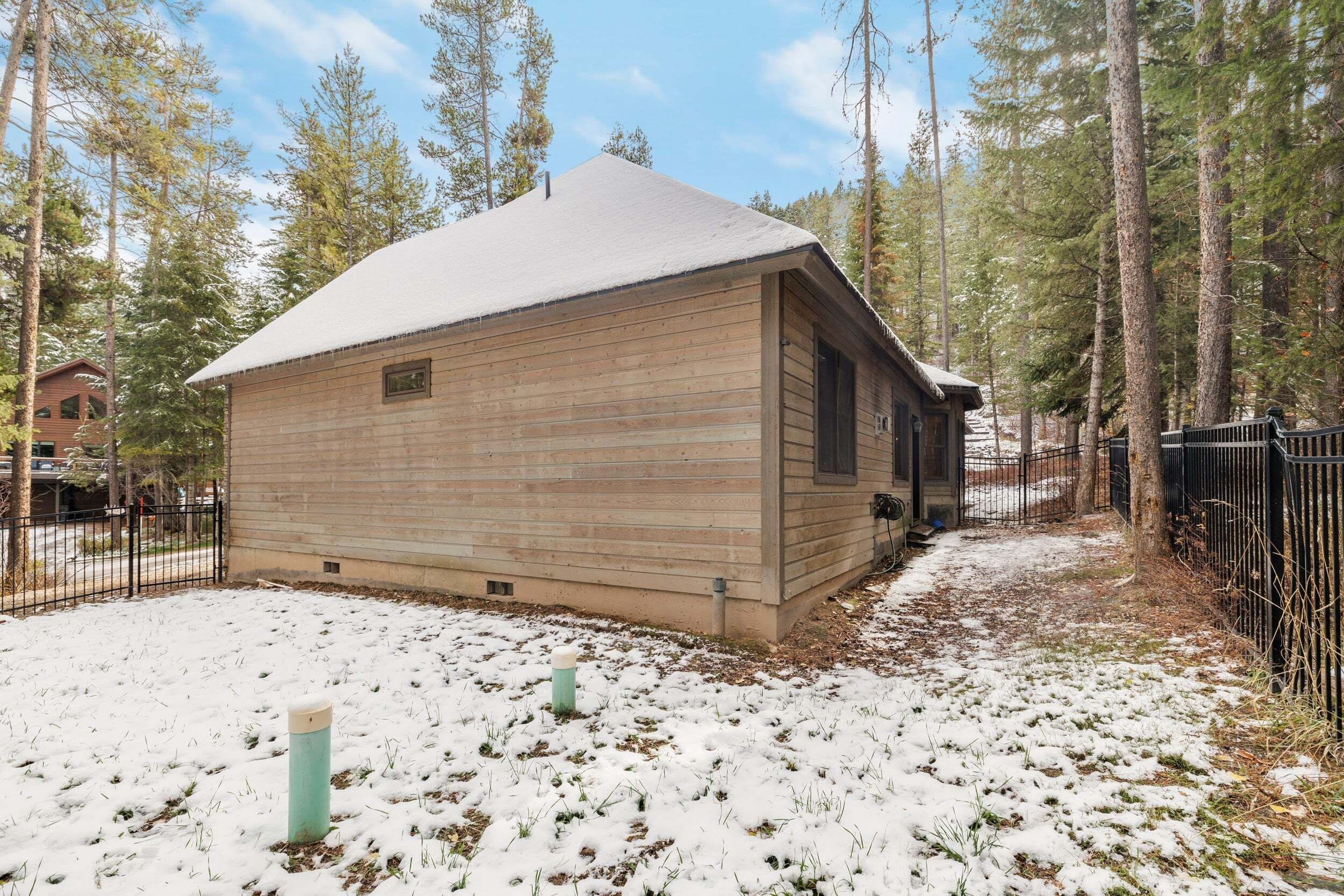 A wooden cabin with a snow-covered roof, surrounded by a forest and a metal fence.