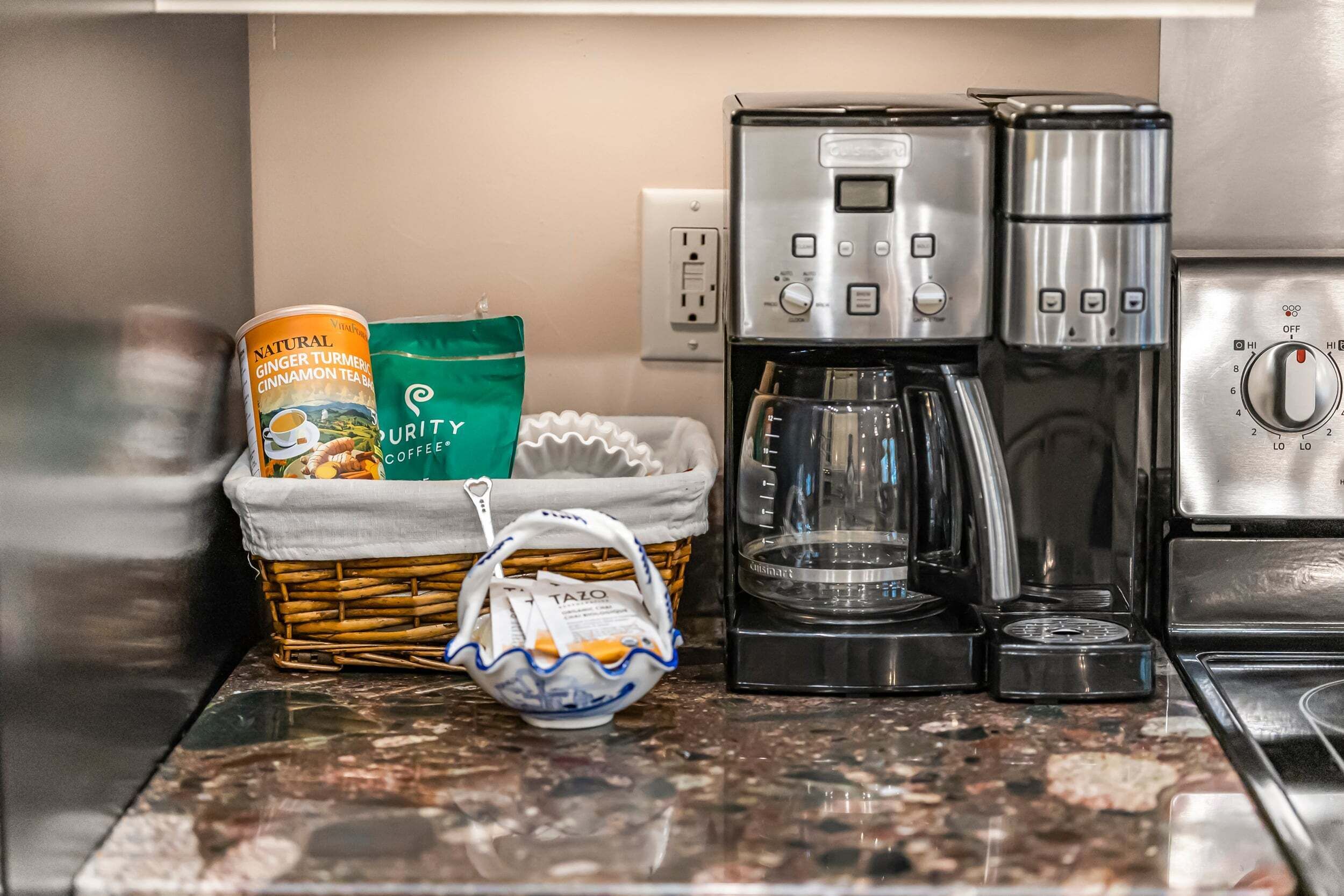 Una encimera de cocina con una cafetera, una cesta que contiene una bolsita de té y una de café, y una pequeña bolsa con un diseño azul y blanco.