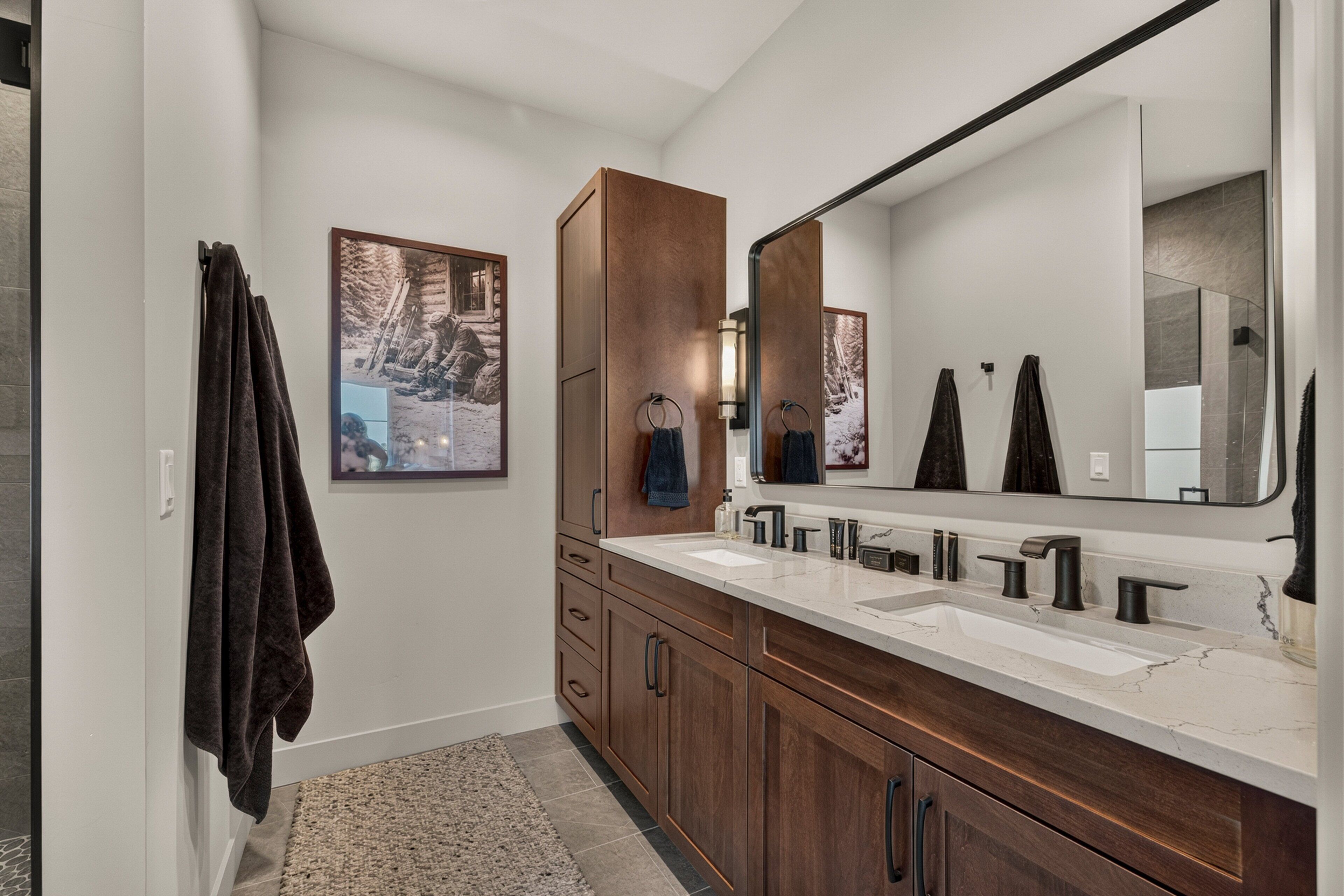 A modern bathroom with a double sink vanity, wooden cabinets, a large mirror, and two framed pictures on the wall.