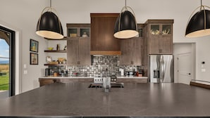 A modern kitchen with a large island, dark countertops, and pendant lighting.