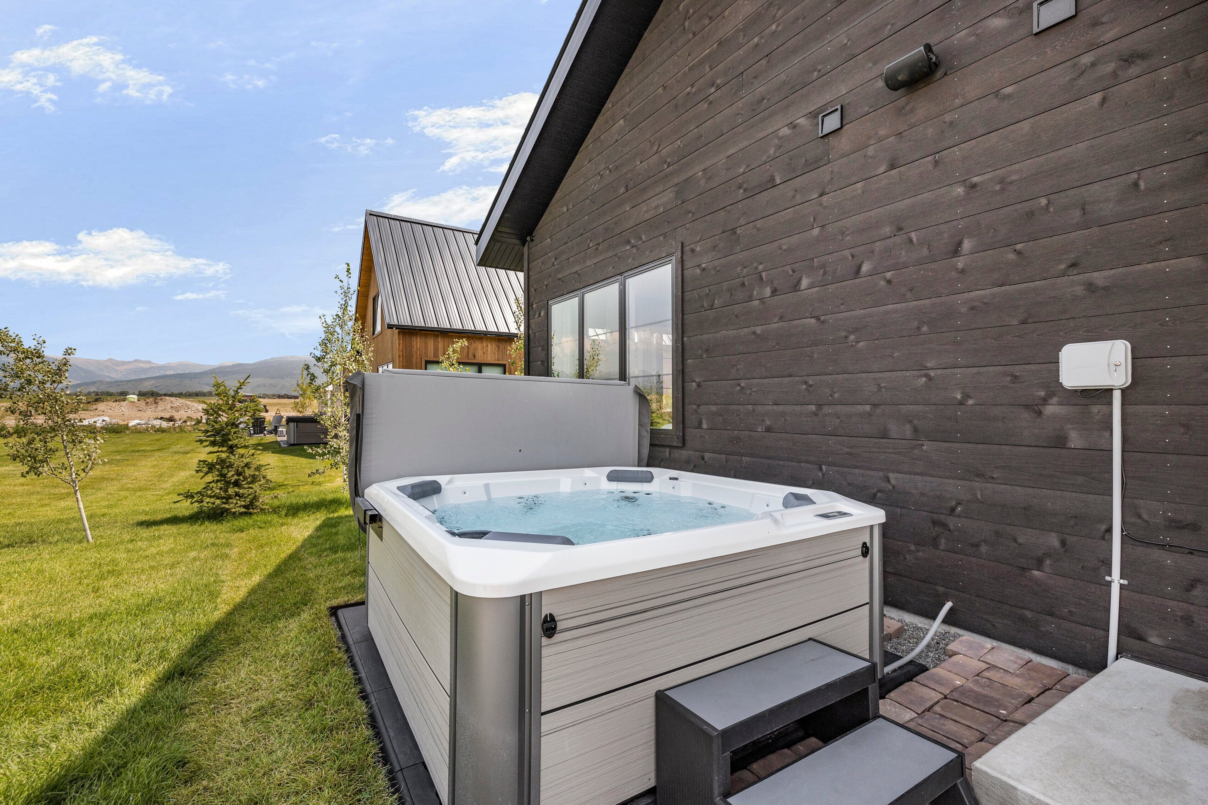 Un jacuzzi al aire libre frente a un edificio moderno, con césped y montañas al fondo.