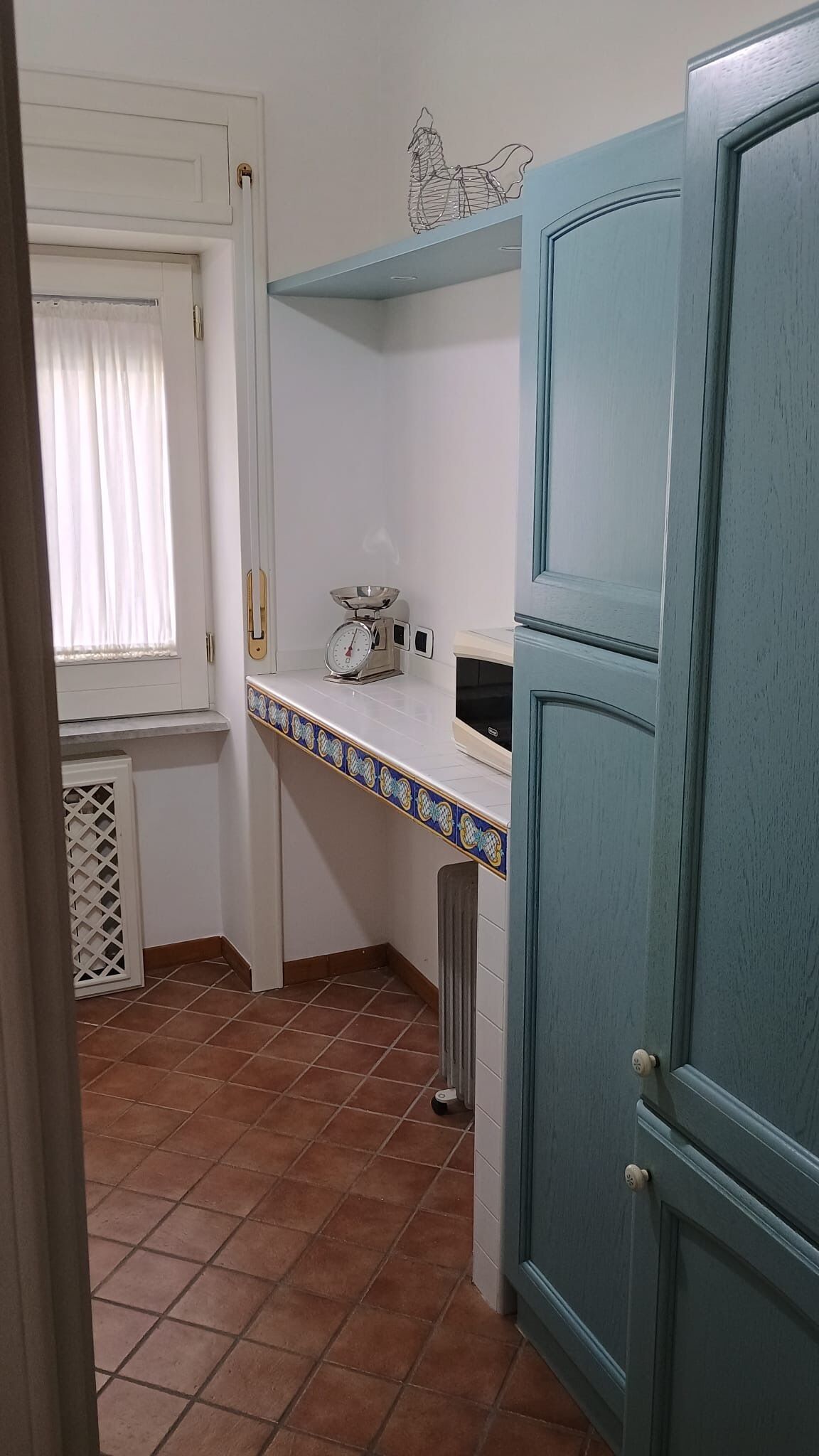 Une cuisine avec un sol carrelé, une fenêtre avec des rideaux, une balance et un meuble bleu.