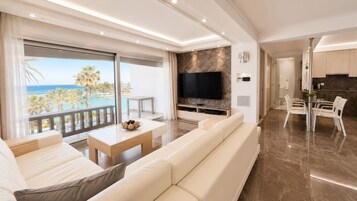 Un salón moderno con un televisor grande, un balcón con vistas al mar y un comedor con sillas blancas.