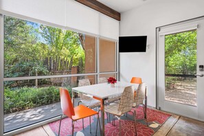 Zona pranzo con tavolo e sedie, televisione a parete e ampie finestre che offrono vista sul verde circostante.