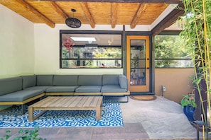 A covered patio with a wooden ceiling, a gray sectional sofa, a wooden coffee table, patterned rugs, and a large window with a view of greenery. - Santa Fe Oasis With Three Primary Suites and Garden Nooks, Can't Miss (Santa Fe)