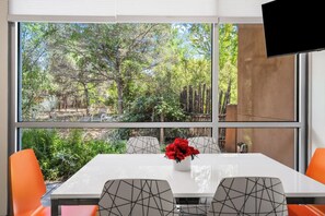 A room with a large window overlooking a forested area, a white table with a vase of flowers, and modern chairs. - Santa Fe Oasis With Three Primary Suites and Garden Nooks, Can't Miss (Santa Fe)
