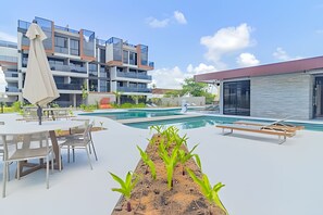 A modern outdoor pool area with lounge chairs and a table, surrounded by a multi-story building with large windows. - Cais Eco Residence beira-mar Muro Alto (Ipojuca)