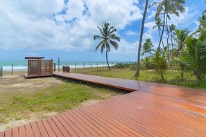 A wooden deck leading to a beach with a wooden structure, palm trees, and a clear sky. - Cais Eco Residence beira-mar Muro Alto (Ipojuca)