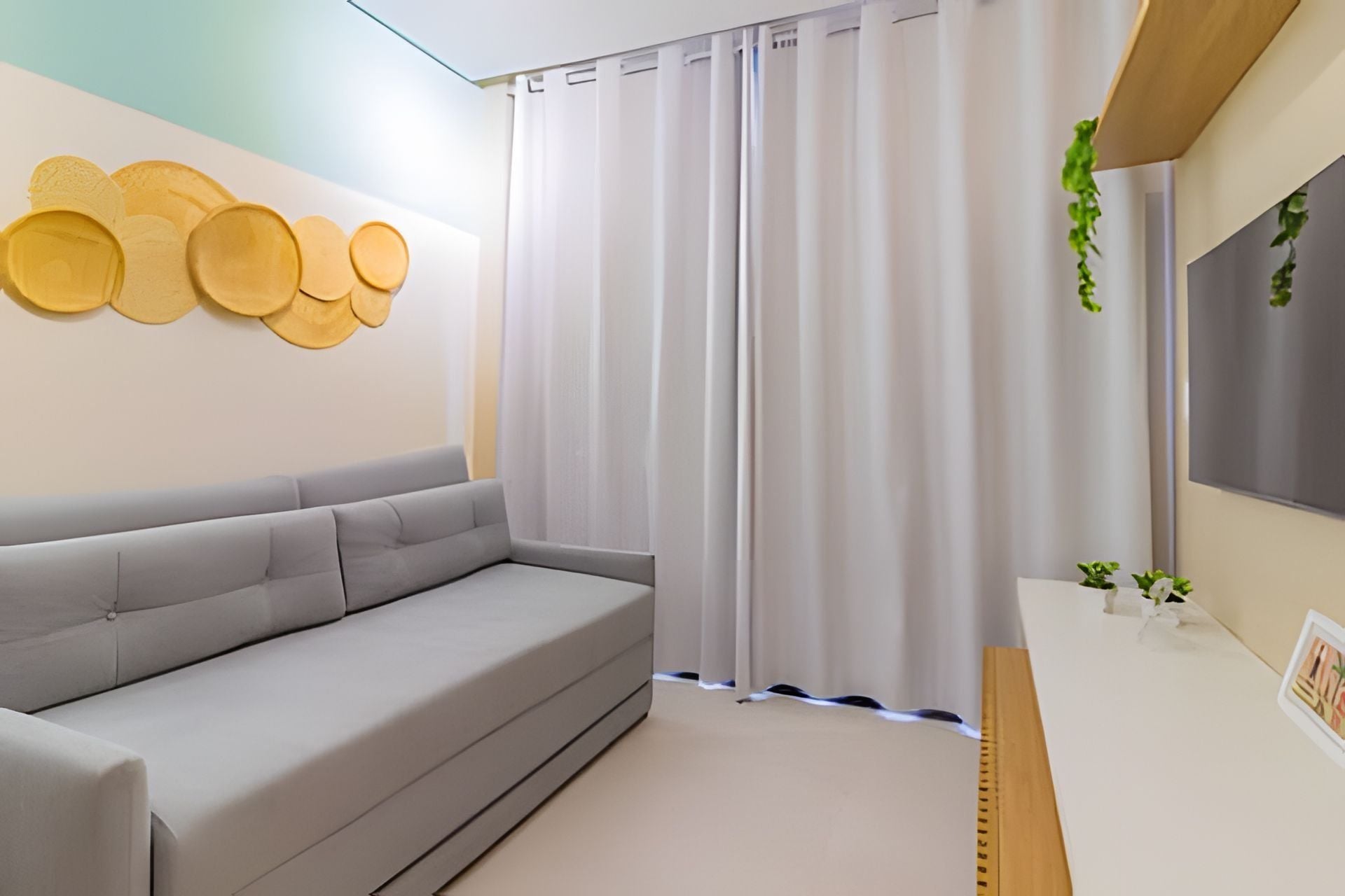 A modern living room with a grey sofa, a wall-mounted decorative panel, a window with white curtains, and a small plant on the windowsill.