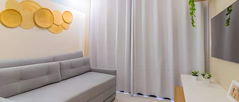 A modern living room with a grey sofa, a wall-mounted decorative panel, a window with white curtains, and a small plant on the windowsill.