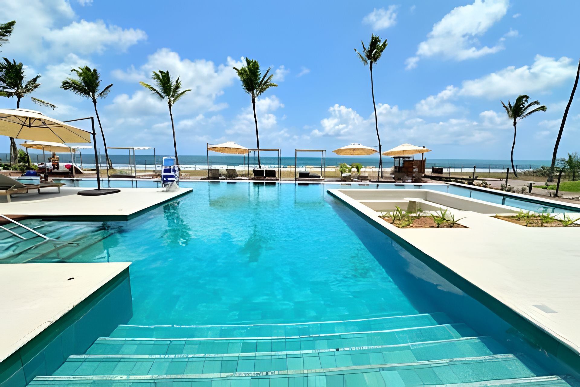 Une piscine à l’eau d’un bleu cristallin, entourée de chaises longues et de parasols, avec des palmiers et une vue sur l’océan.
