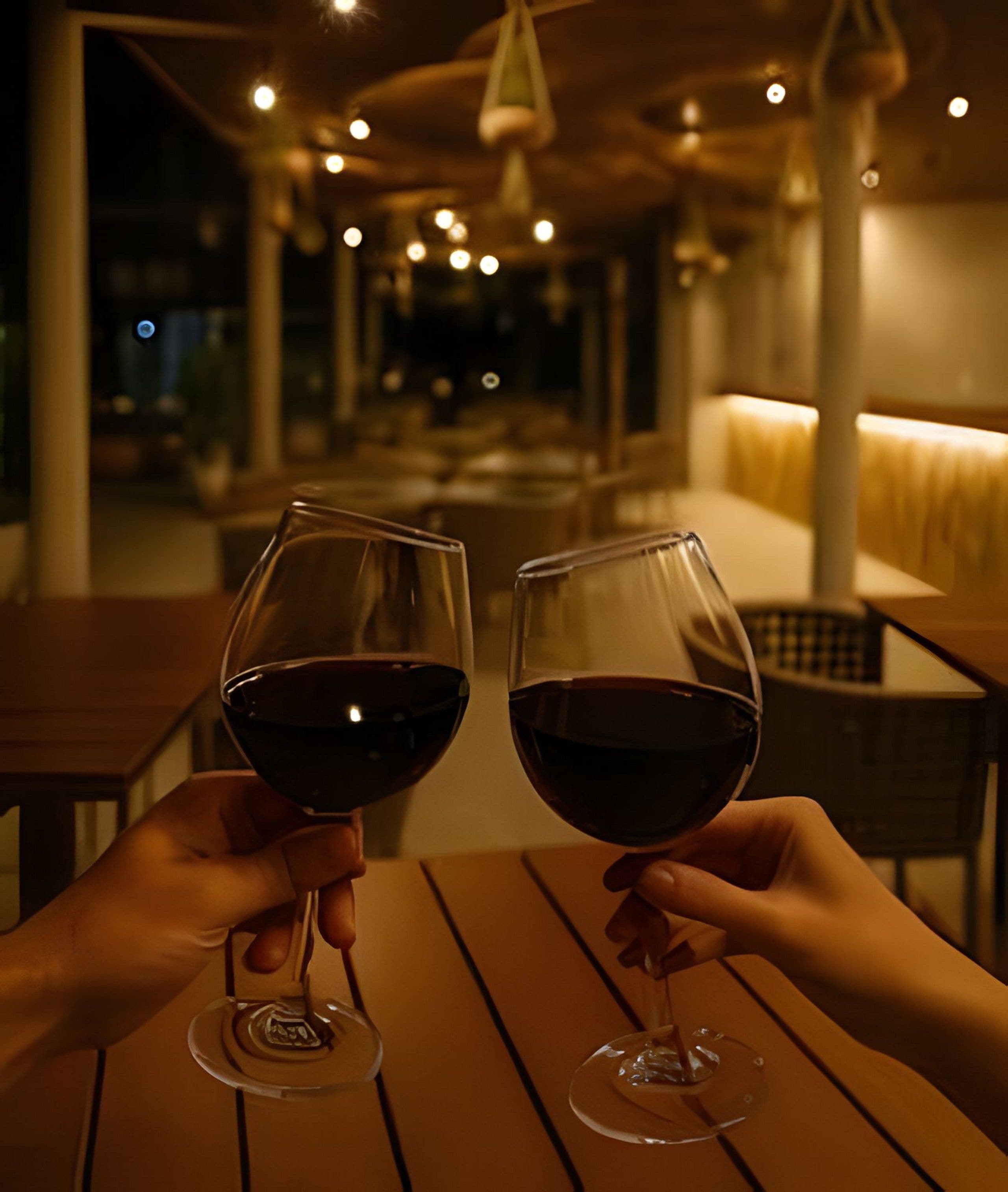 Two hands holding wine glasses at a wooden table in a dimly lit restaurant with a view of the interior.
