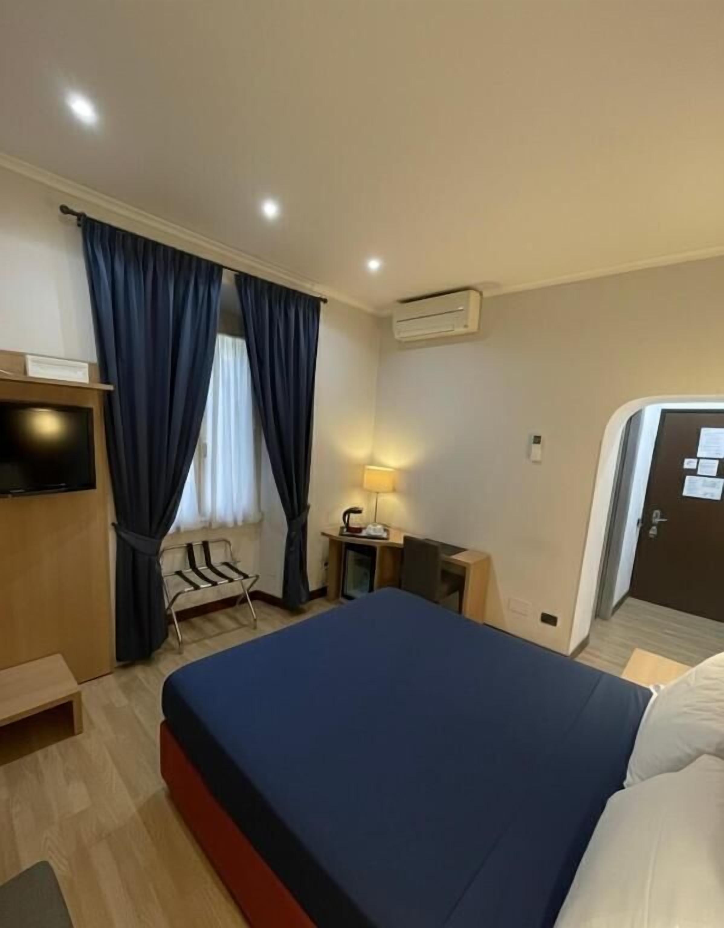 A hotel room with a bed, a desk, a chair, a TV, and an air conditioning unit.